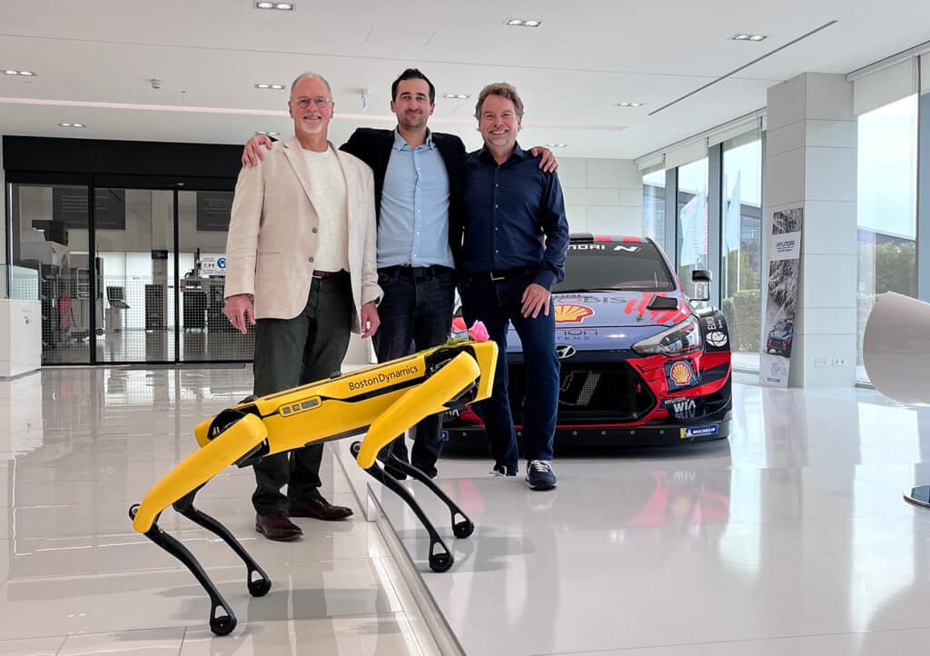 CEO Robert Playter, VP of Sales Philip Archambault, and CSO Marc Theermann pose with Spot in from of a Hyundai at the co-located Hyundai WIA and Boston Dynamics office