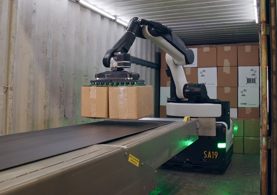 The Stretch robot is grasping 2 brown boxes simultaneously with the suction gripper at the end of an industrial arm, hovering over a conveyor belt inside a container.