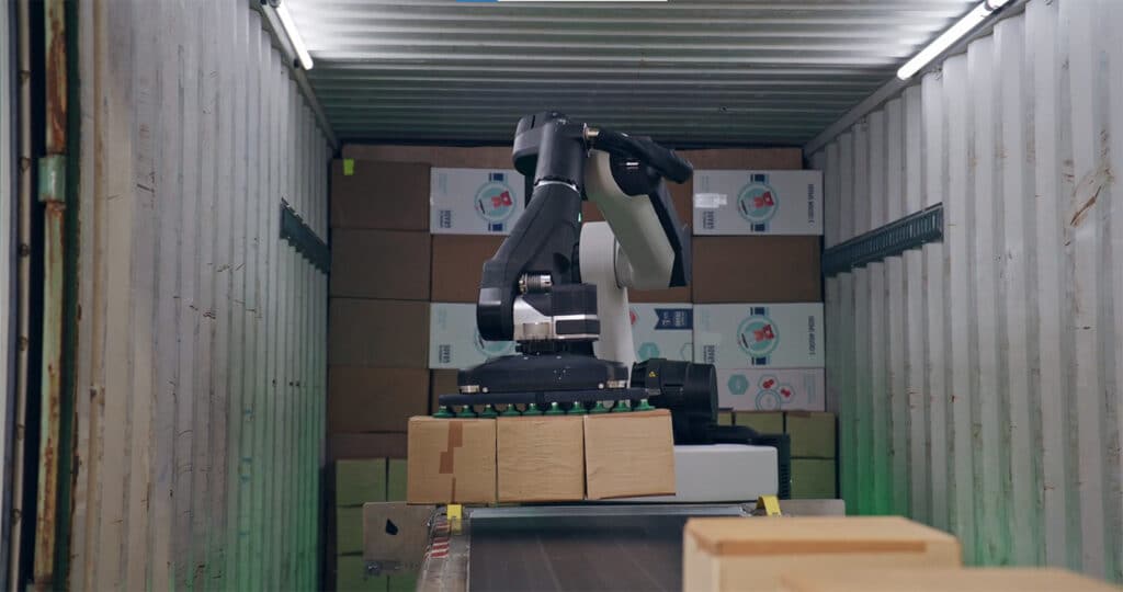 The Stretch robot is grasping 3 brown boxes simultaneously with the suction gripper at the end of an industrial arm, hovering over a conveyor belt inside a container.