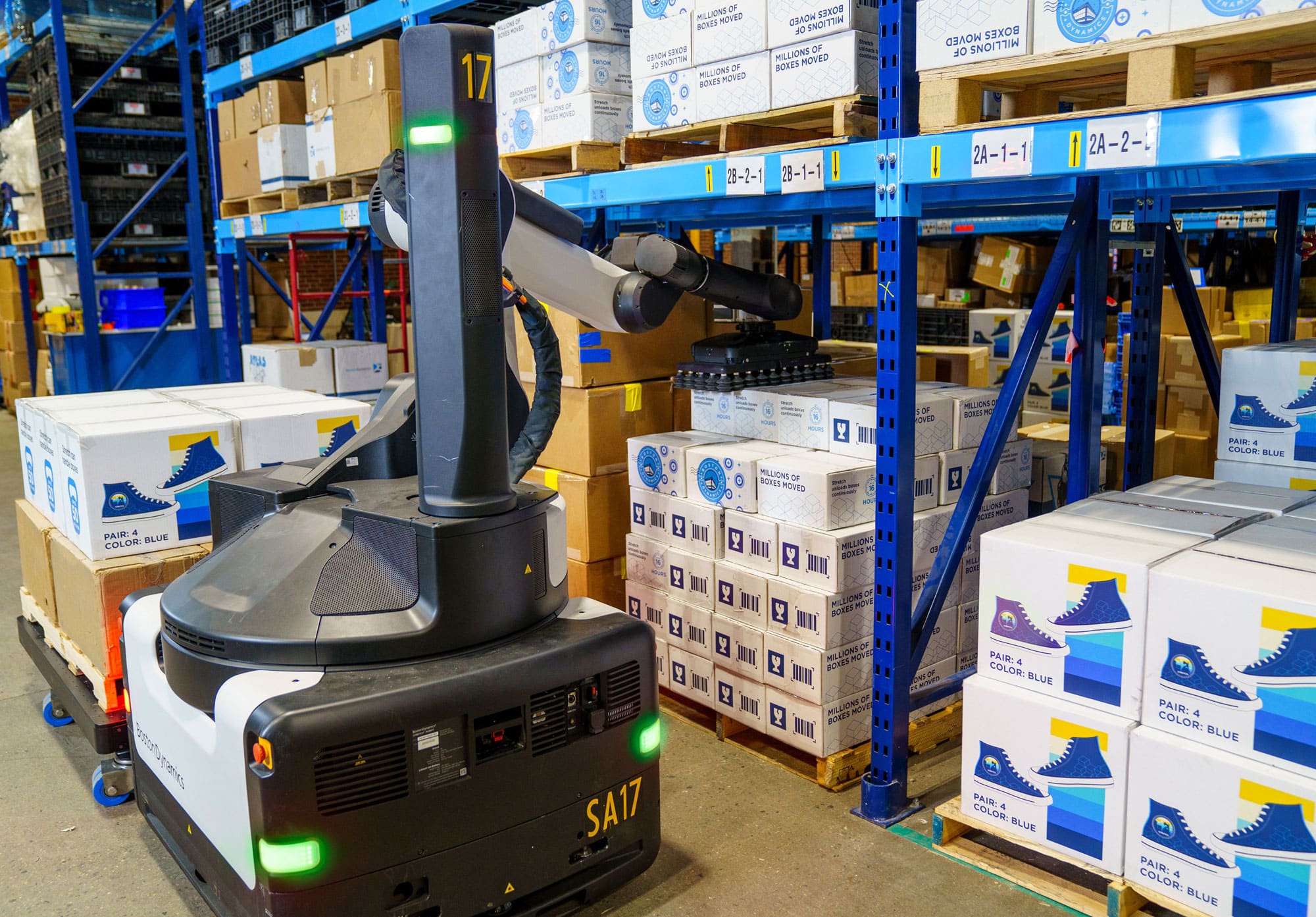 A robot with an arm and gripper grasps a box under racking in a warehouse.
