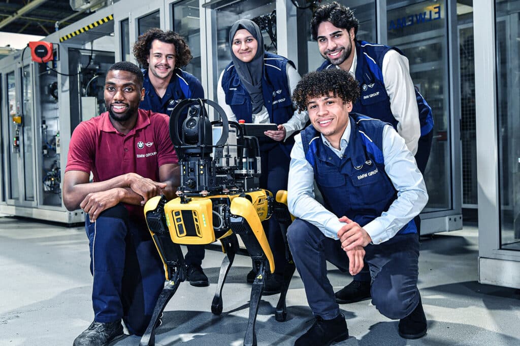 A group of 5 young engineers pose with Spot in an industrial facility