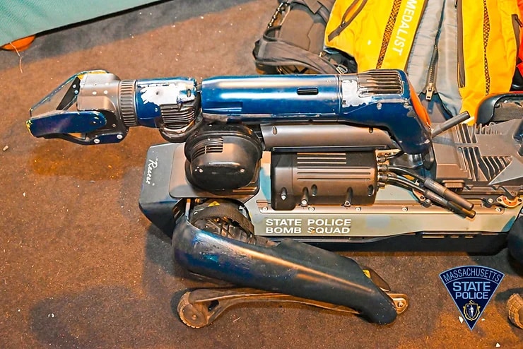 A Spot robot labeled "State Police Bomb Squad" lies on the ground. Several bullet holes are visible on the robot.