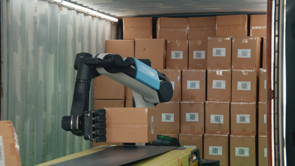 A robotic arm bent over a conveyor grips a cardboard box using its suction gripper, with a wall of boxes behind.