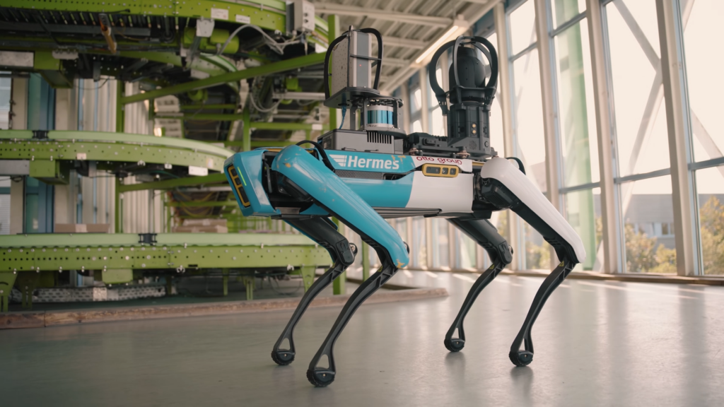 A blue and white quadruped Spot robot stands in front of conveyance systems.