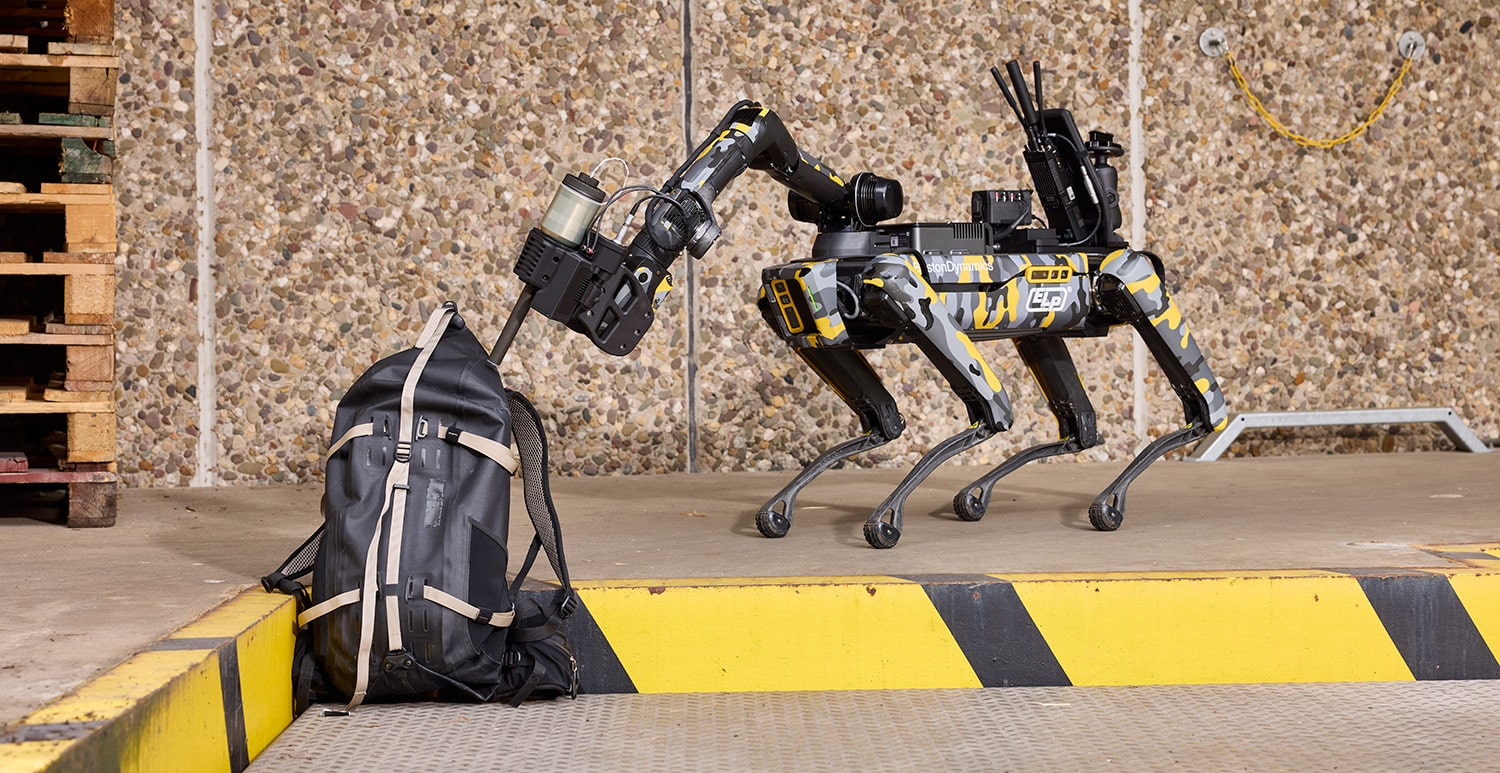 A Spot robot uses its arm to point a disrupter at a bag