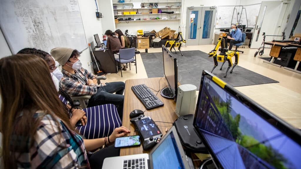 Several group of students work at desks to program two Spot robots in background