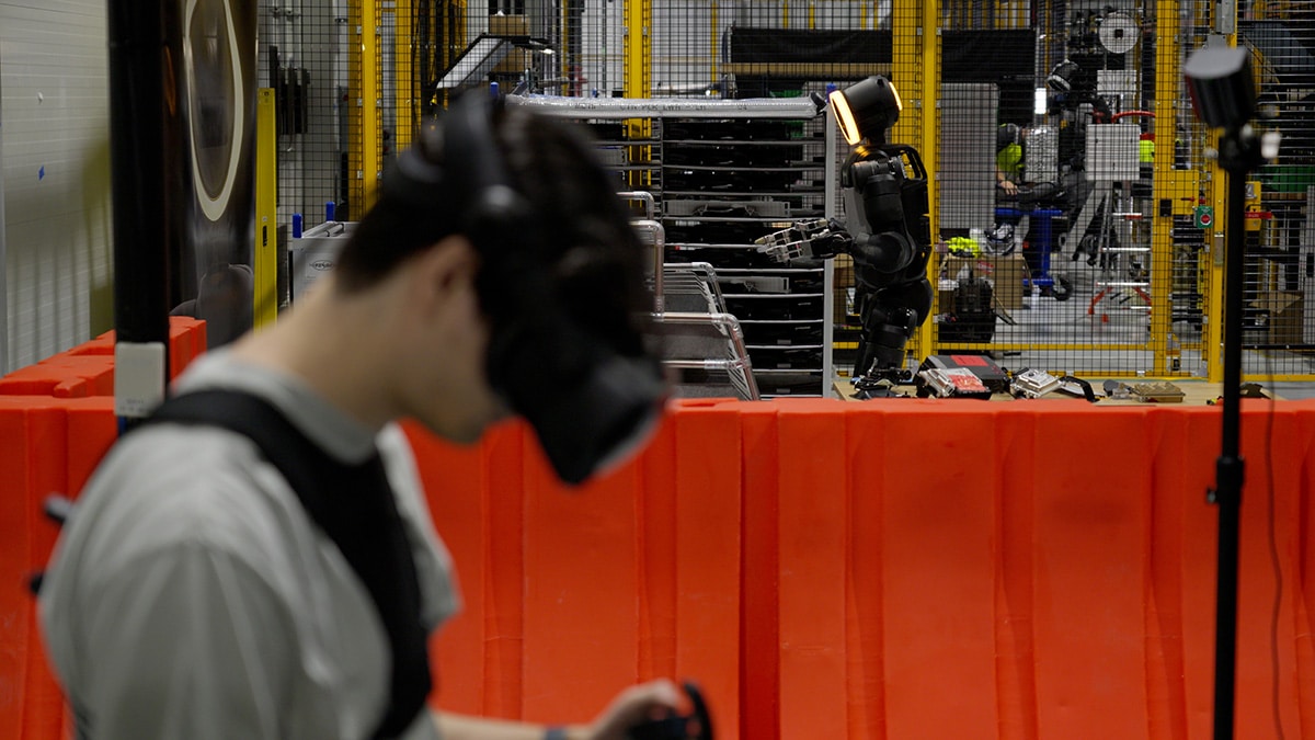 VRヘッドセットを着けた人の奥で、黄色い金網と赤い仕切りの内側にAtlasが立つ(Boston Dynamics 公式リリースより)